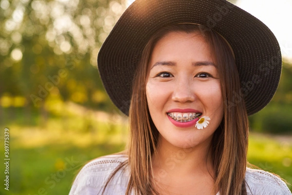 Fototapeta Braces on the teeth, and in the mouth a chamomile flower, a girl of Asian appearance in a white T-shirt and a dark hat, looks at the camera and smiles, blurred background