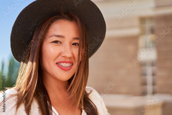 Fototapeta Braces on the teeth, a girl of Asian appearance in a white T-shirt and a dark hat, looking at the camera and smiling, blurred background