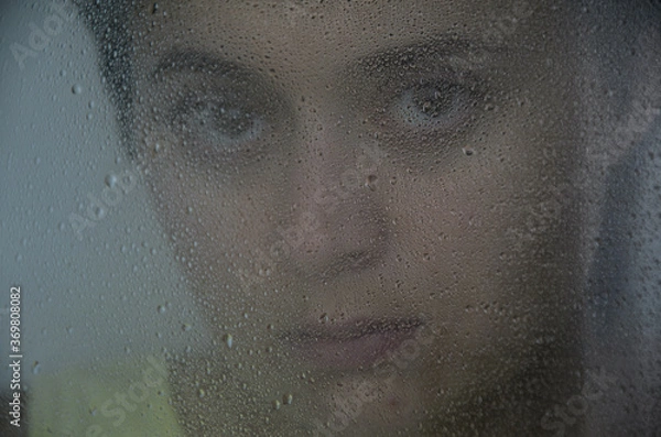 Fototapeta Portrait of a young brunette woman behind glass with rain drops. The concept of depression, blurring about the future, determination. Focus on drops on glass.
