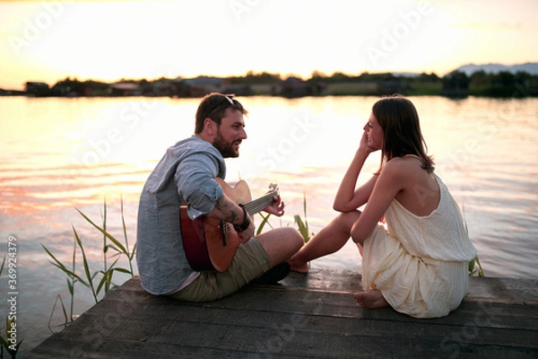 Fototapeta caucasian female enjoying in company of a beardy male playing guitar by the lake at sunset