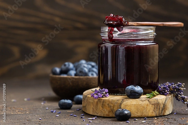 Obraz Glass jar and spoon with delicious blueberry jam with fresh berries, lavender and rosemary on brown table. Healthy food for breakfast.