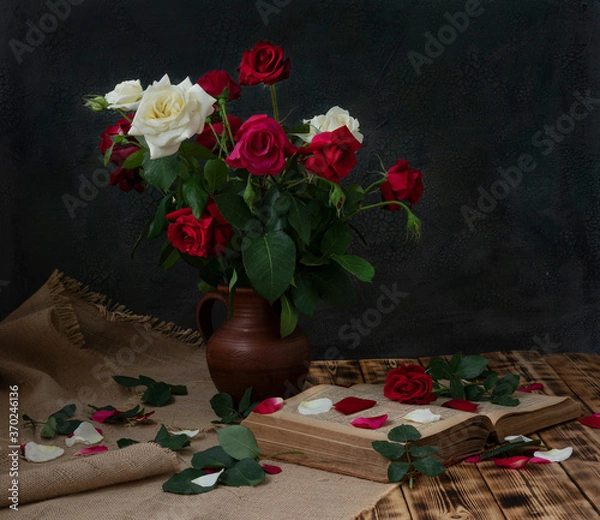 Fototapeta A bouquet of white and red roses stands in a clay jug on a wooden table. An old book is lying nearby.