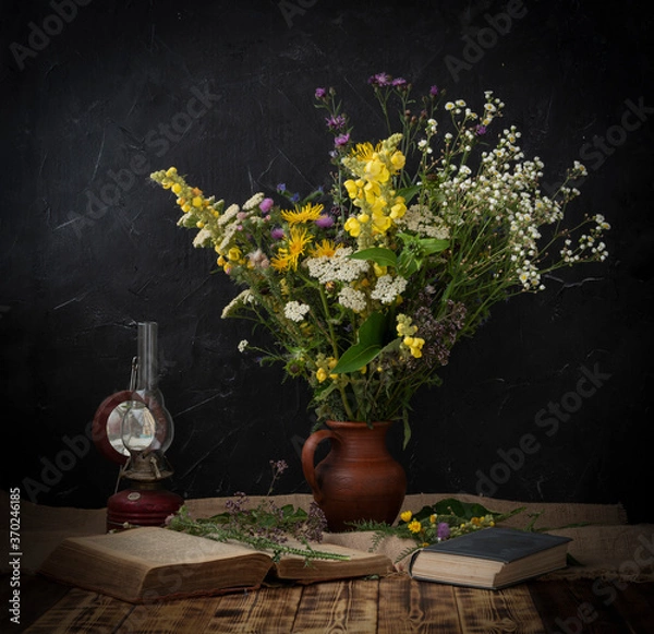 Fototapeta A bouquet of wildflowers is on a wooden table, there is an old lamp next to it and books are lying.