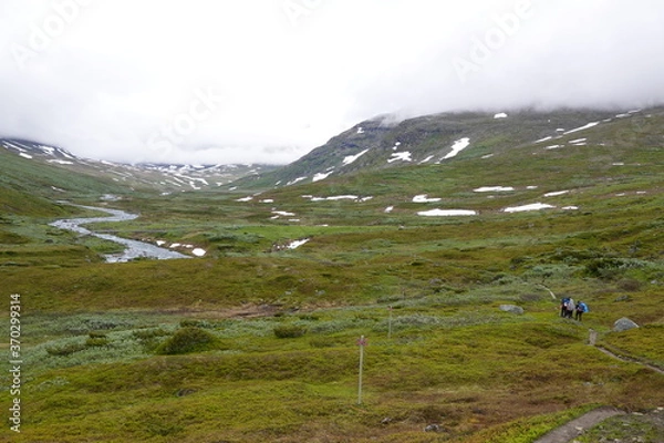 Fototapeta People on an hiking trail in a green valley in Sweden with a river, clouds and some snow