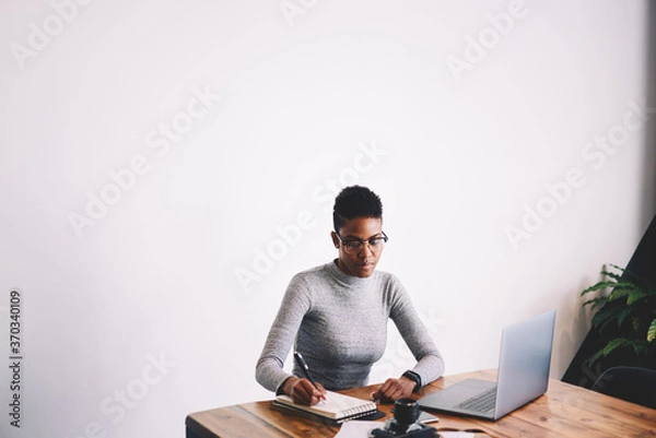 Fototapeta Concentrated Afro-American female designer creating sketch in notebook concentrated on ideas, professional businesswoman in eyewear planning projects making notes sitting on promotional background