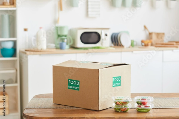 Fototapeta Background image of cardboard box with food standing on wooden table in white kitchen interior, food delivery service, copy space