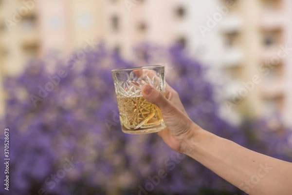 Obraz A hand holds a glass of white wine whisky against the background of houses and a blooming jacaranda tree in Lisbon, Portugal.