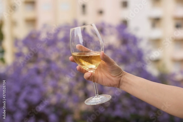 Obraz A hand holds a glass of traditional white green wine against the background of houses and a blooming jacaranda tree in Lisbon, Portugal.
