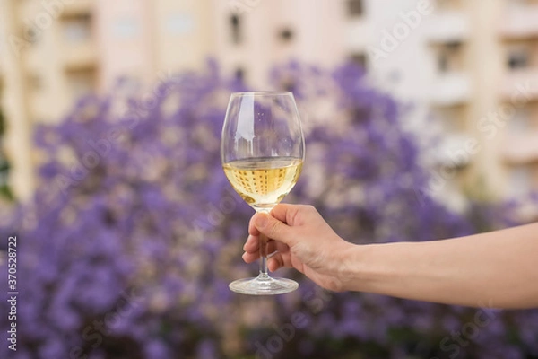 Obraz A hand holds a glass of traditional white green wine against the background of houses and a blooming jacaranda tree in Lisbon, Portugal.