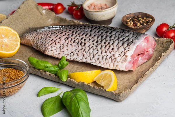 Fototapeta Headless carp carcass, peeled from fish scales, with lemon, cherry tomatoes, pepper and spices on a large clay dish on a gray background