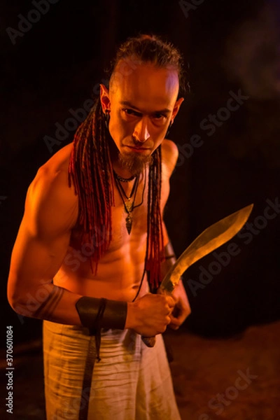 Fototapeta Irdorath group, clip making backstage. The caucasian man with dreadlocks,  with knife in the hand. Viking's nighttime in the forest is lighting by flamed torches.