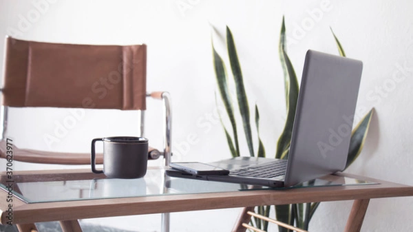 Obraz Trabajar desde el hogar. Plano frontal en un interior iluminado y blanco de una taza negra de cafe junto a una notebook y un teléfono celular en un pequeño escritorio. 