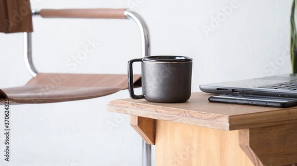 Obraz Trabajar desde el hogar. Plano frontal en un interior iluminado y blanco de una taza negra de cafe junto a una notebook y un teléfono celular en un pequeño escritorio. 