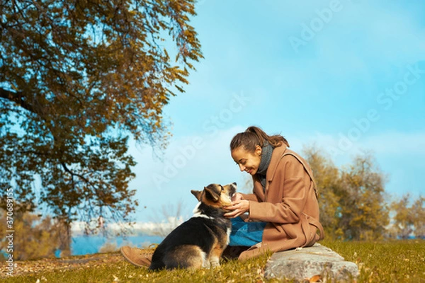 Obraz Jesienne spacery ze zwierzakiem. kobieta w stylowym płaszczu siedzi ze swoim psem Welsh Corgi w jesiennym parku, patrzy na siebie, kontakt wzrokowy ze zwierzakiem. Pojęcie przyjaźni z psem i człowiekiem, portret jesień
