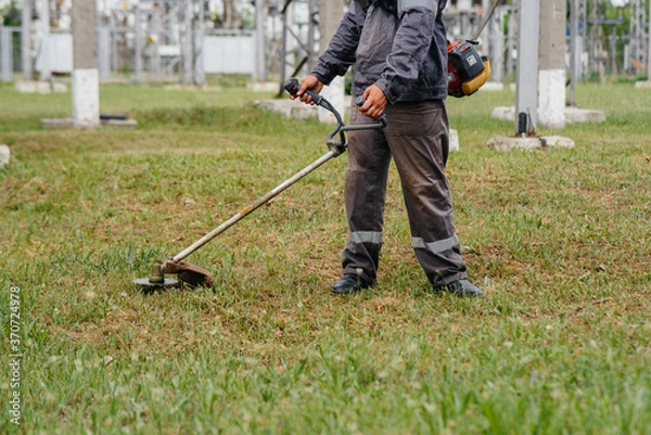 Fototapeta A young man mowing grass on the territory of an electric substation in overalls. Grass cleaning at the enterprise, implementation of fire safety measures