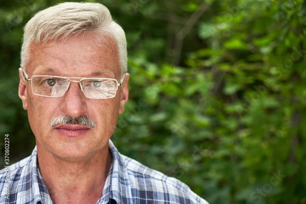 Fototapeta Close-up portrait of handsome gray-haired adult man with mustache and glasses smiling on the background of a summer forest park