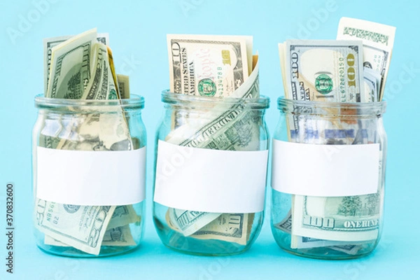 Fototapeta Three Piggy Bank from a Glass Jar for Life. New dollars sticking out of piggy banks on a blue background. The banks have white sheets for inscriptions. Mockup. Copy space. Economy concept