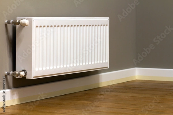 Obraz White metal heating radiator mounted on gray wall inside a room.