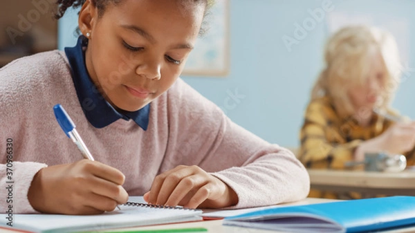 Fototapeta In Elementary School Classroom Brilliant Black Girl Writes in Exercise Notebook, Taking Test and Writing Exam. Junior Classroom with Group of Bright Children Working Diligently and Learning New Stuff