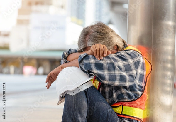 Fototapeta Sad fired worker with safety helmet hat sitting on street after being dismissed..Engineer unemployed from company  sitting sad outside office in city background. Man fired from job.