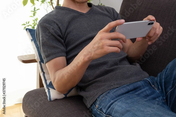 Fototapeta ソファでくつろぎながらスマホで動画を見ている男性