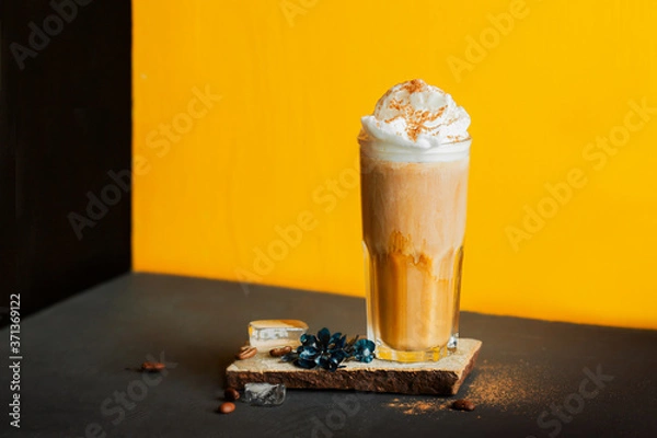 Fototapeta cold black coffee with whipped milk foam and spices in high glass on stand of natural stone, coffee beans and ice cubes on black table and yellow background