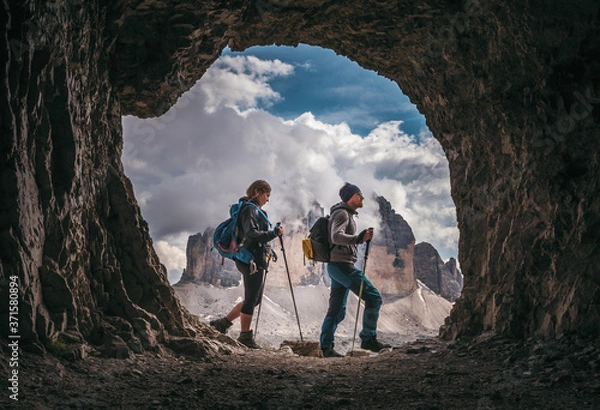 Obraz Couple of hikers walking, with Tre Cime di Levarado peaks on background, view from a cave window, in Dolomites, Italy. 