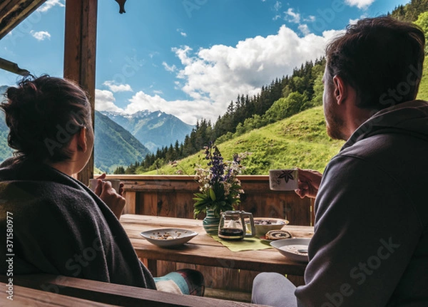 Obraz Couple sitting outside of a mountain hut, eating breakfast and enjoying the scenery