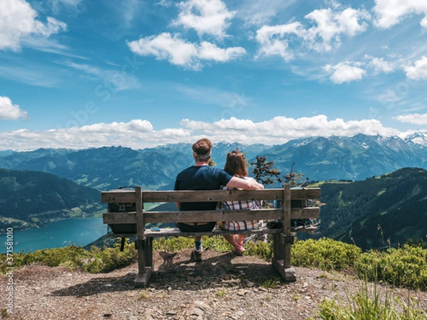 Obraz Young couple sitting on bench on top of a mountain enjoying the scenery of Zell am Zee panorama in Austria
