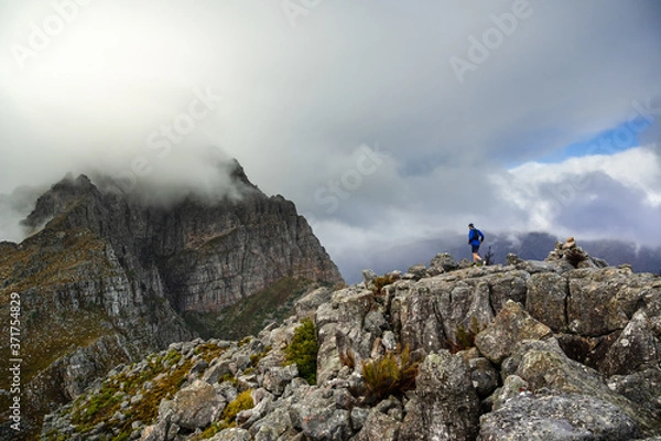 Fototapeta A trail runner running along a steep rocky ridge in the high mountains while heading off on an adventure to climb a big mountain