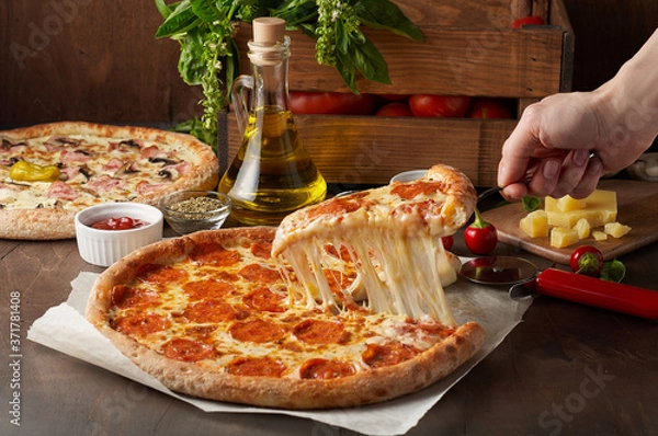 Fototapeta Female hand hold slice of hot pizza with melting cheese and chorizo on rustic wooden table with ingredients