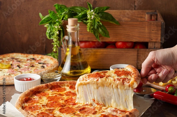 Fototapeta Female hand hold slice of hot pizza with melting cheese and chorizo on rustic wooden table with ingredients
