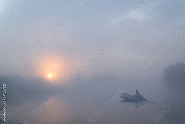 Obraz Tranquil morning landscape panorama at sunrise. A fisherman on a rubber boat on the river in heavy fog. Dawn illuminates and makes the clouds colorful.