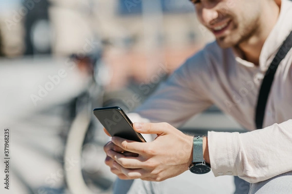 Fototapeta Online in city. Smiling attractive guy in shirt sits on street and looks at smartphone