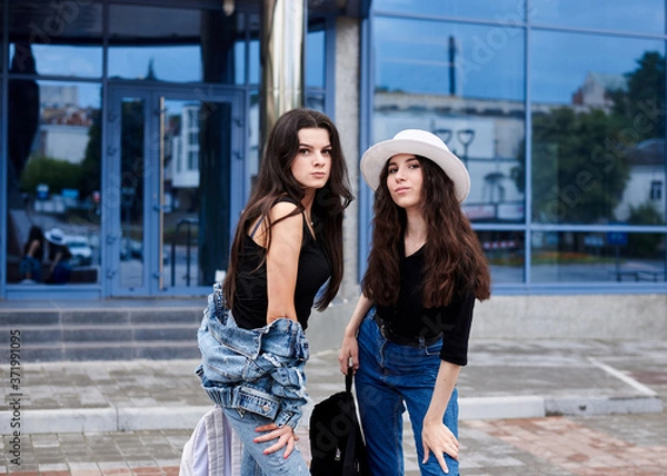 Fototapeta Two young brunette girls, wearing casual jeans attire, standing in front of blue modern glass building. Girlfriends traveling in city, holding backpacks, sightseeing. Active lifestyle concept.