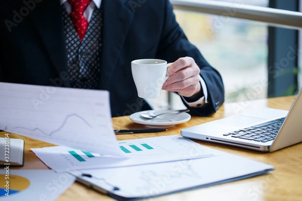 Obraz Adult male businessman working on a new project and looking at stock growth charts. Sits at a large window at the table. Looks at the laptop screen and drinks coffee.