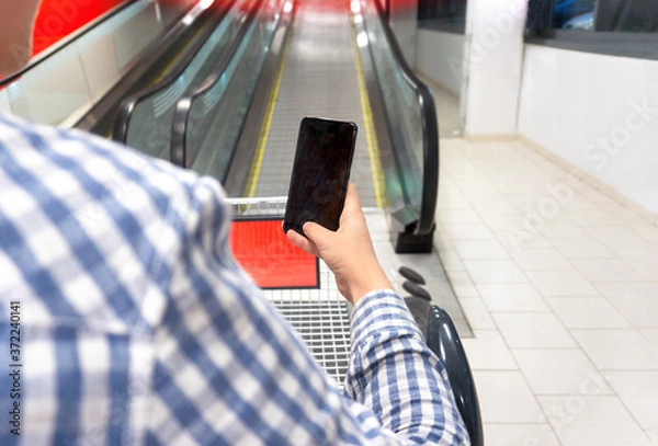 Obraz Hand holding mobile phone on escalator background. Concept of online ordering of goods with delivery via applications for mobile phone