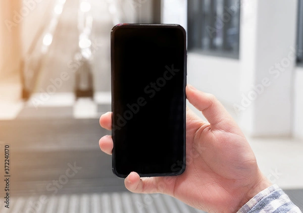 Obraz Hand holding mobile phone on escalator background. Concept of online ordering of goods with delivery via applications for mobile phone