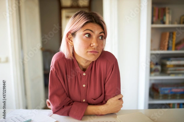 Obraz Portrait of sad student girl with nose ring and pink bod haircut staying home having bored facial expression, looking away. Confused young Caucasian female posing indoors, sitting at table