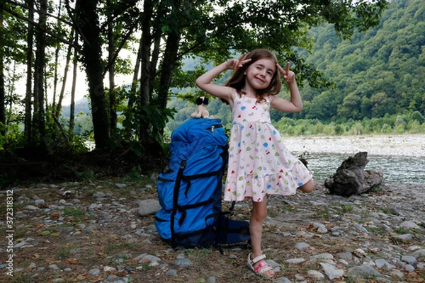 Obraz A large backpack for an adult stands next to a little girl against the backdrop of trees and a river, on a sunny day.