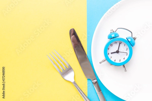 Fototapeta Blue alarm clock, fork, knife and empty plate on colored paper background. Intermittent fasting concept