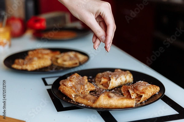 Fototapeta Woman's hand decorates pancakes with a chocolate, fruits kiwi and banana. Black plate, white table