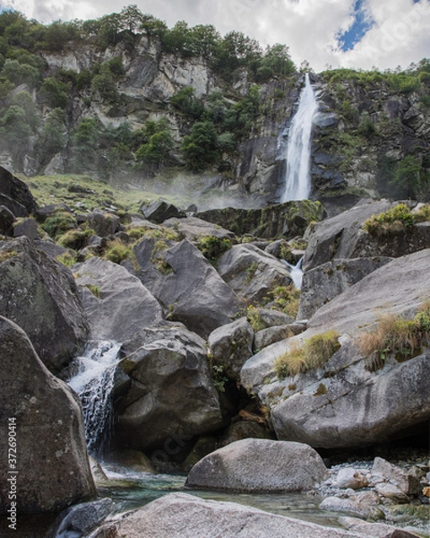 Fototapeta Wasserfall in den Alpen vor Felsen bei wolkigem Himmel in der Sonne
