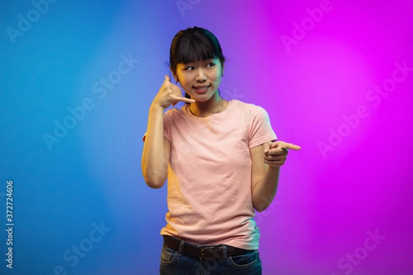 Fototapeta Need you to call. Asian young woman's portrait on gradient studio background in neon. Beautiful female model in casual style. Concept of human emotions, facial expression, youth, sales, ad.