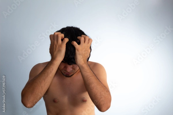 Obraz Young naked Hispanic man with wavy hair and shaved beard on a white background, covering his face with his hands tightly