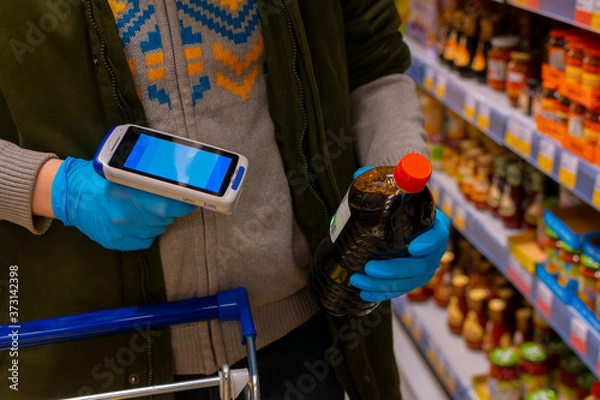 Fototapeta A customer wearing medical protective gloves holds a barcode scanner and scans the product. Cropped shot, no face visible. Self-service in the store.