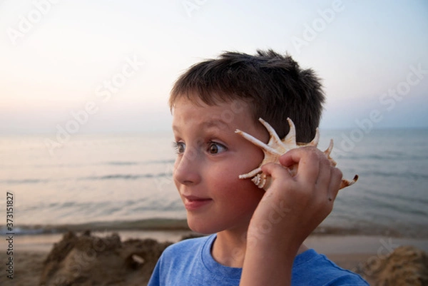 Fototapeta handsome boy with a surprised expression on his face listens to a seashell. The concept of cheerful and joyful emotions.