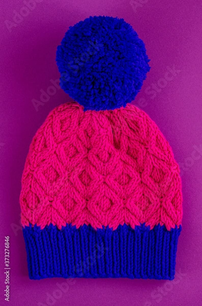 Obraz Beautiful two color handknitted hat with pompon on purple background. Top view