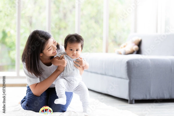 Fototapeta Happy little baby learning to walk with mother help in living room. Baby taking his first steps with mother's help and support with love.