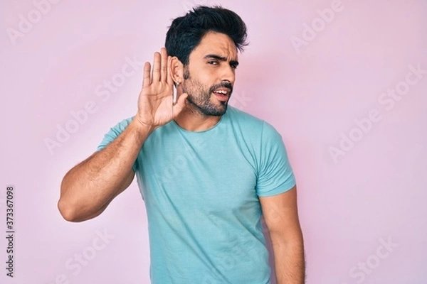 Fototapeta Handsome hispanic man with beard wearing casual clothes smiling with hand over ear listening an hearing to rumor or gossip. deafness concept.